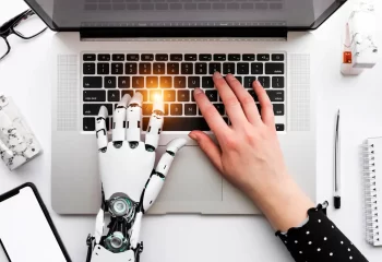 Imagem de uma m&atilde;o humana e uma m&atilde;o rob&oacute;tica digitando em um laptop, representando a integra&ccedil;&atilde;o entre humanos e intelig&ecirc;ncia artificial no uso de tecnologia.