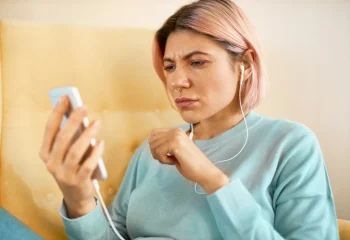Mulher jovem com cabelo rosa usando fones de ouvido e olhando com express&atilde;o preocupada para o celular
