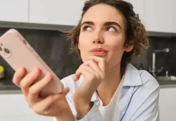 Mulher pensativa segurando um smartphone em uma cozinha moderna, refletindo sobre suas op&ccedil;&otilde;es.