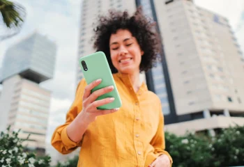 Mulher sorrindo usando camisa amarela segurando smartphone com capa verde em &aacute;rea urbana com pr&eacute;dios ao fundo