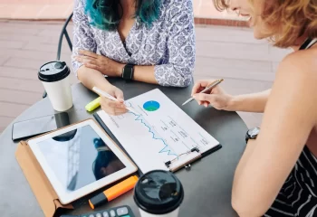 Duas mulheres analisando gr&aacute;ficos financeiros e dados em papel com tablet, smartphone e caf&eacute; em mesa redonda