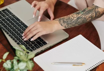 Pessoa trabalhando em um laptop com um bra&ccedil;o tatuado em uma mesa de madeira, com caderno e caneta ao lado, em ambiente de escrit&oacute;rio ou estudo.