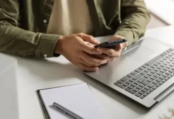 Uma pessoa utilizando um smartphone enquanto est&aacute; em frente a um laptop em um ambiente de trabalho, com um bloco de anota&ccedil;&otilde;es e caneta ao lado.